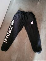 Black Air Jordan sweatpants laid flat on floor showing white lettering down one leg and red and white logo patch