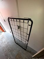 Metal cot frame propped against a wall with a wire grid base and black metal bars.