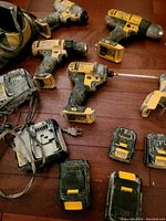 Photo showing four DeWalt cordless drills with battery packs attached, two battery chargers with cords, four batteries, and one flashlight, all showing signs of heavy use and dirt.