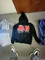 Photo showing a black hoodie with 'JESUS' printed in red dripping letters on the front, laid flat on the floor among other clothing.
