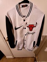 White and black varsity jacket hanging on hanger showing front with Chicago Bulls logo and Nike swoosh, button closure, black and white striped collar, cuffs and hem.