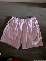 Photo of pink Nike shorts laid flat on a gray surface, showing front side with elastic waistband and white Nike swoosh logo on left leg.