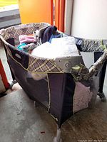 Full view of the baby playpen with fabric and mesh sides and metal legs, showing some soft items inside which are not included.