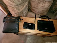 Photo showing three purses on a wooden surface in front of a green chair: a black leather crossbody purse with zippers, a small crocodile-embossed wristlet, and a structured handbag with a top handle.