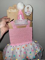 Pink 1st birthday baby outfit displayed on packaging with pink crochet top, polka dot tulle skirt, and matching party hat with pom pom.
