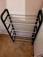 Shoe rack with five shelves made of metal rods and black plastic side frames, shown standing on carpeted floor against white wall.