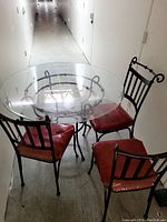Round glass table with black metal decorative base paired with three black metal framed chairs with red cushions in hallway.