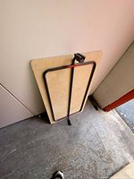 Photo of a rectangular folding tabletop leaning against a wall. The table top is light-colored and has black metal folding legs attached underneath, secured with a red strap.