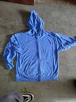Full view of blue TNA towel fabric hoodie with hood and zipper laid flat on floor.