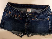 Front view of dark blue denim True Religion women's jean shorts showing button closure, front pockets with rivets, and frayed hems.