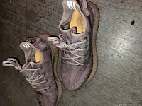 Pair of brown Adidas Yeezy Boost 350 V2 sneakers on floor, showing side and top views with visible reflective heel stripes and ribbed gum soles.