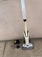 Front view of cordless Hoover vacuum in white with black accents, standing upright on concrete floor with charging cable beside it.