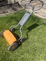 Side angle of push lawn mower showing orange cylindrical blade housing and gray handles with control knobs.
