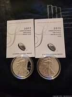Two US Mint Certificates of Authenticity (2015 and 2017) alongside two American Silver Eagle coins in plastic capsules displayed on dark fabric