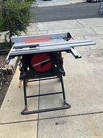 Front angle showing aluminum table surface, red circular saw blade, adjustable fence, and tubular metal stand with wheels and wooden handles