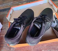 Pair of black Nike golf shoes displayed in an open orange shoebox on a wooden surface.