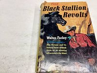 Front cover of The Black Stallion Revolts book showing artwork of a black stallion and rider with cowboy hat. Dust jacket is torn and worn.