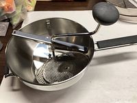 Photo of a stainless steel food mill with crank handle and black grip, showing perforated disc inside bowl