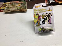 Image shows unopened Hot Wheels The Beatles Yellow Submarine Cockney Cab die-cast car in original packaging placed on a white surface with other items in background.