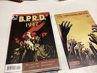 Two B.P.R.D. 1947 comics and The Walking Dead comic laid flat on a white surface