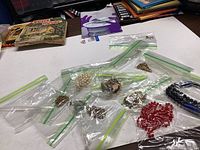 Overview of various jewelry pieces in individual plastic bags on a white surface showing different chain types and colors including pearl-like beads, gold-tone chains, and colored beads.