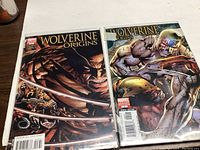Two Wolverine Origins comic books on a wood surface, one with Wolverine in a close-up pose with claws extended, the other showing Wolverine in battle with a cyborg-like character.