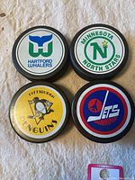 Four NHL hockey pucks showing large team logos for Hartford Whalers, Minnesota North Stars, Pittsburgh Penguins, and Winnipeg Jets