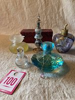 Five vintage perfume bottles arranged on beige cloth. Included are clear faceted glass bottle, pale yellow flower-shaped bottle, blue faceted bottle with pendant, tall detailed bottle with decorative stopper, and purple heart-shaped bottle with gold pump.