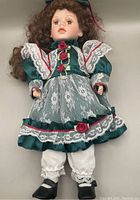 Brown-eyed doll in dark teal dress with lace and red roses, showing bisque head, forearms, and lower legs.