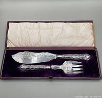 Boxed antique fish serving set including knife and fork with ornate silver handles inside a velvet-lined box with satin underside lid.