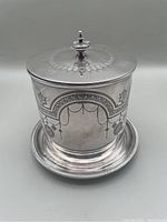 Front view of the antique silver plated biscuit barrel showcasing ornate engraved decoration and knob on lid.