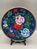Front view of cloisonné plate showing colorful floral design with pink, red, blue flowers, green foliage, and a butterfly on a blue background with gold rim, on a black stand.