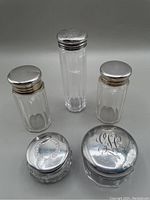 Overview of 5 dresser jars showing clear crystal bodies with sterling silver lids varying in size and shape, 2 lids gold washed, 4 lids monogrammed. Worn condition on 3 lids.