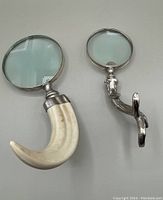 Two magnifying glasses laid flat: one with a horn-shaped white cellulose plastic handle and metal frame, the other with a silver metal ergonomic handle