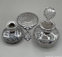 Two small perfume bottles with silver overlay and a small silver plate handheld mirror. The bottles show intricate floral and vine silver overlay designs on clear glass. The mirror is oval-shaped with decorative silver plate backing and a dent on the back.