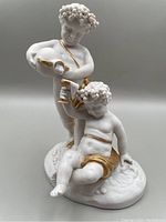 Front view of Spanish biscuit porcelain cherubs figurine showing standing cherub pouring liquid and seated cherub holding cup, gold accents on garments and grapevine hair detail
