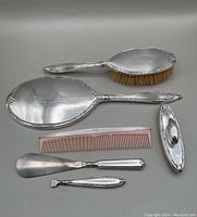 Full view of 6 piece antique sterling silver dresser set including mirror, brush, comb, shoe fitter, nail buffer, and nail file handle on white background.
