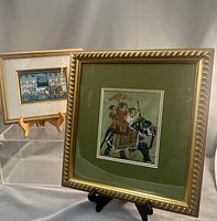 Both framed paintings displayed with stands. One depicts royal figure on elephant with umbrella bearer and attendants. The other shows procession with elephants and architectural backdrop.