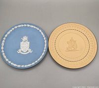 Top view of two Wedgwood Jasperware plates side by side on a surface. Left is blue with white coat of arms, right is cane yellow with cherubs and dark blue border.