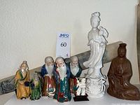 Photo showing eight Asian figurines on a table including small glazed ceramic figures with green glaze and crazing, three elderly men figurines in colored robes, a tall white porcelain standing figure holding a bird, and a seated brown figure. A rolled Thai rubbing scroll is in the background.
