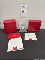 Photo of the Baccarat crystal clock placed between the original red box and instruction booklets, showing clock front, box with Baccarat logo, and manuals.