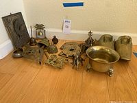 Wide shot showing the full lot of brass items including the stirrup shoes, bells, trivet, brass bowl, wall hanging, and various figurines arranged on a wooden floor against a cream wall.