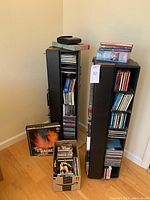 Two black CD racks side by side on floor, one with top stack of CDs and accessories, cardboard box of discs in front