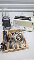Overall lot view showing the NutriBullet blender, Cuisinart toaster, flatware in organizer tray, and kitchen utensils.