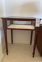 Front view of a wooden-framed glass display case side table with tapered legs, showing minor nicks on corners.