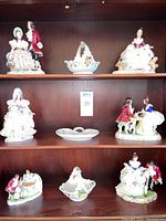 Full shelving unit displaying 6 Dresden porcelain lace figurines and 3 porcelain dishes in excellent condition, arranged on three shelves.