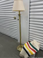 Brass floor lamp with fabric shade beside folded wool blanket with colorful stripes.