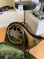 Overview of French horn in open green velvet-lined case with stool in background