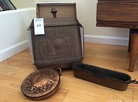 Photo of embossed metal lavabo with coat of arms, pierced brass overlay lantern, and copper fish kettle with handle on wood floor
