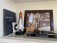 Overall view of the space collection showing the model NASA Endeavour space shuttle with external fuel tank, framed vintage satellite photo collage with 'Dynamics Explorer' title, Draper Laboratory book, and Daphne Satellite toy in display box